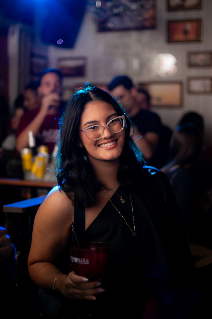 Smiling woman with glasses holding drink at lively indoor bar setting.