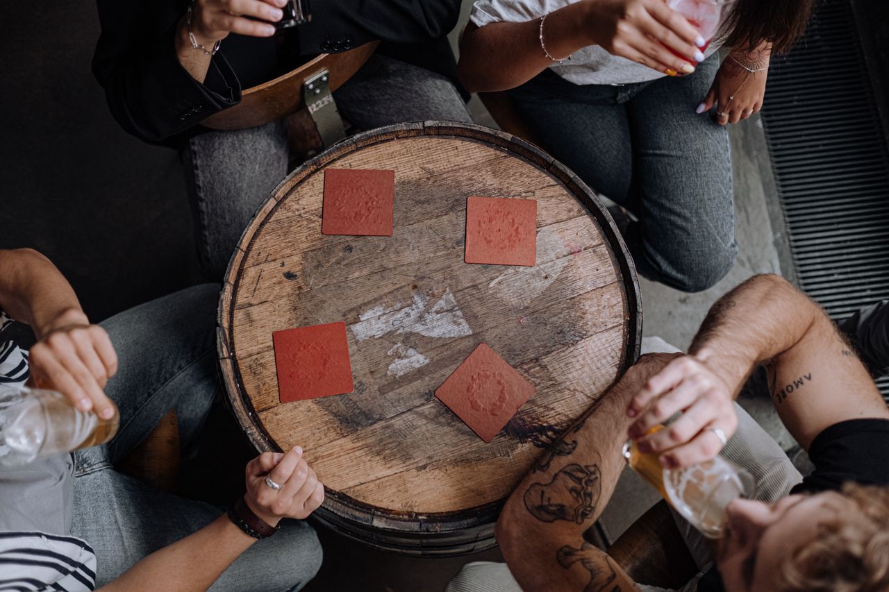 A group of friends gathered around a rustic barrel table with drinks and coasters, photographed from above.